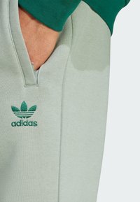 Trening Adidas verde deschis, cu guler cu fermoar și logo brodat verde, având o textură netedă și un design minimalist.