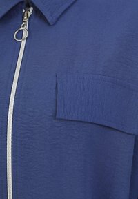 Tummansininen takki, jossa on vetoketjullinen etuosa, kärjellinen kaulus ja paikka-tasku. Materiaalissa on hienovarainen pinta ja pehmeä viimeistely.