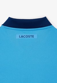 Ljusblå polo med en mörkblå krage. En rektangulär etikett med ordet "LACOSTE" är sydd på baksidan.