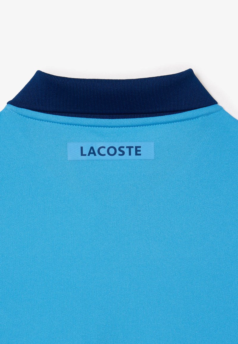 Polo bleu clair avec un col bleu foncé. Une étiquette rectangulaire avec le mot "LACOSTE" est cousue à l'arrière.