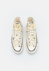 Sapatilhas altas em lona branca com respingos de tinta amarela e preta. Apresentam atacadores brancos, biqueira de borracha e detalhes em preto.