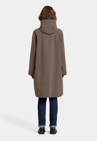 Pitkä ruskea vedenpitävä takki hupulla, jossa on suora leikkaus, sivutaskut ja napituksia, pukeutuneena tummiin farkkuihin ja mustiin saappaisiin.