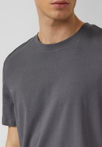 Szary bawełniany t-shirt z okrągłym dekoltem, krótkimi rękawami i subtelnym wytłoczonym logo na piersi. Gładka faktura i klasyczne dopasowanie.