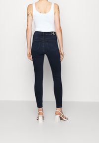 Helle Skinny Jeans aus einem dunklen Denim mit mittelhoher Taille, ausgestattet mit Gesäßtaschen und einem kleinen schwarzen Logo am Bund. Kombiniert mit einem weißen Oberteil und hohen Schuhen.