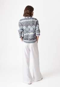 Adīta džemperis ar ziliem un baltiem rakstiem, kas satur dažādas ģeometriskas formas, saskaņots ar baltām lina platām biksēm.