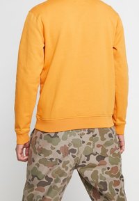 Oranje katoenen sweatshirt met een ronde halslijn, ribgebreide manchetten, gedragen met beige broek met camouflagepatroon en achterzakken.