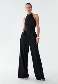Melnā halter stilā jumpsuit ar pieguļošu bodi un plaša kāju dizainu. Tekstūrēta auduma, plecēta vidukļa un minimāliskiem zelta aksesuāriem.