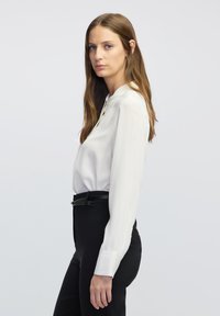 Weißes Langarmbluse mit rundem Ausschnitt und gefältelten Manschetten, kombiniert mit figurbetonten schwarzen Hosen. Schlichtes, sauberes Design und glattes Material.