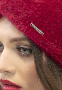 Cappello rosso peloso con un'etichetta metallica che riporta la scritta "Vivienne" nella parte anteriore, indossato in modo obliquo, coprendo parzialmente i capelli e la fronte del modello.