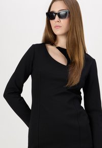 Fekete ruha hosszú ujjal, kivágott nyakrésszel és arany kiegészítőkkel. A modellen fekete napszemüveg van.