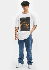 Hvid oversized t-shirt med et grafisk tryk af en kronet figur. Båret med lysblå denimbukser og hvide sneakers.
