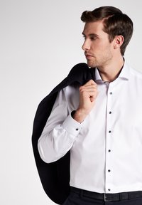 Weißes Hemd mit spitzen Kragen, blauen Knöpfen und französischen Manschetten, kombiniert mit einem dunklen Sakko über einer Schulter, schwarzem Gürtel und taillierten Hosen.