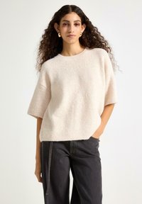 Lysebeige kortærmet sweater i blød, struktureret strik med rund hals. Parret med mørke, løstsiddende jeans.