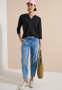 Femme portant un haut à manches longues noir, un jean bleu clair, des baskets blanches, une casquette rose, tenant un sac vert olive, debout contre un mur beige.