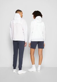 Jachete albe, ușoare, cu glugă și manșete elastice, cu o textură netedă; purtate peste pantaloni bleumarin și pantaloni scurți, cu teniși albi.