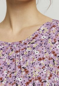 Lilla tunika med blomsterprint i gul, hvid og mørkerød. Har plisseret udskæring og blød stofstruktur for komfort.
