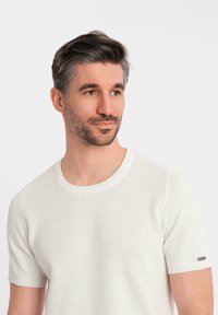 Suéter de punto blanco de manga corta con un patrón texturizado y un escote redondo. Presenta un pequeño acento metálico en la manga izquierda.