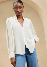 Blusa bianca con scollatura a V e chiusura a bottoni. Caratterizzata da maniche a sbuffo e pizzo sul colletto e sui polsini. Abbinata a jeans blu.