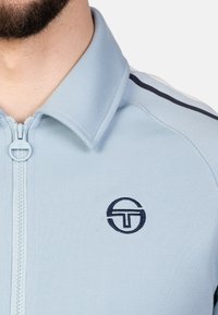 Helles blaues Zip-Jackett mit Kragen, das marineblaue Streifen auf den Schultern und ein rundes marineblaues Logo auf der linken Brustfläche aufweist.