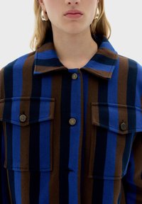 Camicia con bottoni a righe marroni, blu e nere. Presenta un colletto, due tasche sul petto e dettagli in metallo con bottoni. Tessuto texturizzato.