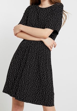 Vrouw draagt een zwarte jurk met korte mouwen met klein wit stippenpatroon en elastische taille, armen gekruist over haar torso.