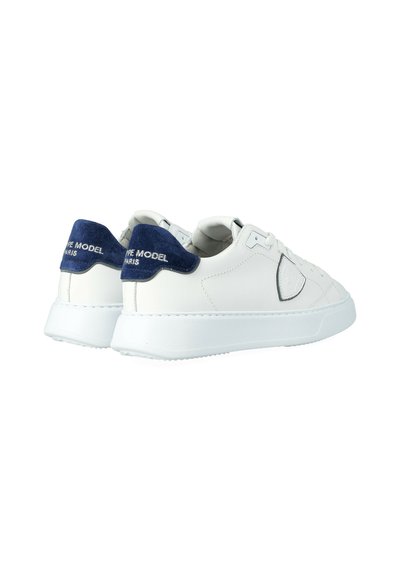 Een paar witte low-top sneakers met navy blauwe hieltabs en de tekst "TYPE MODEL PARIS", gestikte details en witte rubberen zolen.