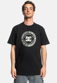 Černé bavlněné tričko s velkým kruhovým grafickým motivem v krémové barvě a textem. Krátké rukávy a kulatý výstřih, s volným střihem.