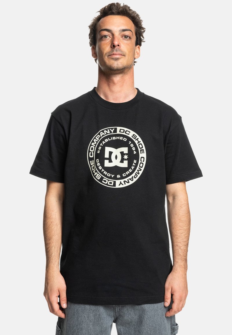 Černé bavlněné tričko s velkým kruhovým grafickým motivem v krémové barvě a textem. Krátké rukávy a kulatý výstřih, s volným střihem.