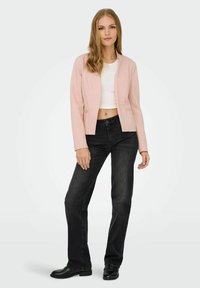 Femme debout portant un blazer rose clair, un haut court blanc, un jean droit noir et des mocassins noirs, sur un fond uni.