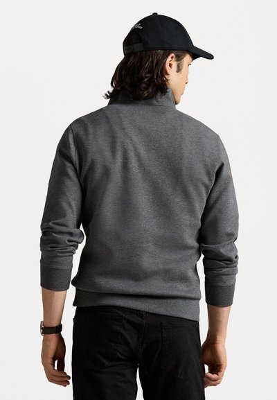 Grijze sweatshirt met een hoge kraag, geribbelde manchetten en onderrand. Draagt zwarte broek, gezien van achteren. Zwarte pet zichtbaar op het hoofd.