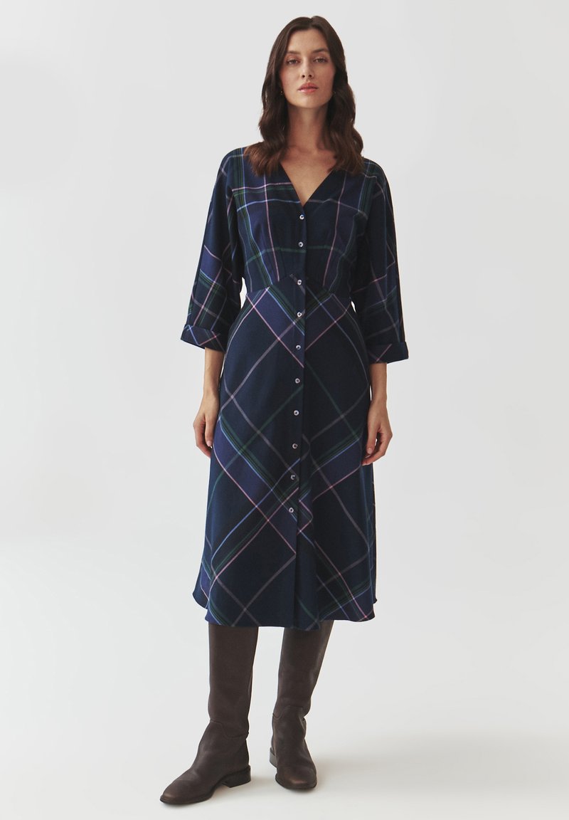 Robe à carreaux bleu marine avec un décolleté en V, des boutons sur le devant et des manches ¾. Elle présente des rayures vertes, roses et blanches dans un motif entrecroisé, d'une longueur mi-mollet.