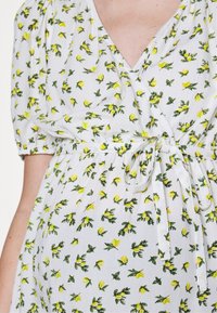 Robe portefeuille blanche avec imprimé floral jaune, manches courtes bouffantes et ceinture nouée à la taille, portée par une personne vue du cou jusqu'aux hanches.