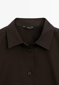 Chemise marron foncé à col avec des boutons noirs et une étiquette de marque visible au niveau du col.