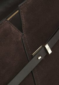 Tummanruskea mokkanahkainen käsilaukku, jossa musta nahkahihna ja kullanvärinen lukko, jossa on kohokuvioitu logo; yksityiskohtaiset ompeleet ja vetoketjun reuna näkyvissä.