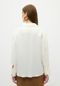 Blusa bianca in seta con colletto, polsini abbottonati con dettagli a taglio e orlo ondulato, abbinata a pantaloni marroni.