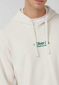 Acercamiento de una persona que lleva una sudadera color crema con el texto "s.Oliver Co. Establecido en 1969" y cordones, sobre una camiseta blanca.