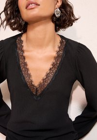 Žena v černém topu s dlouhými rukávy a hlubokým véčkovým výstřihem lemovaným průsvitnou květinovou krajkou a stříbrných kroucených náušnicích ve tvaru kruhu.