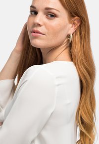 Top bianco a maniche lunghe con una texture liscia, caratterizzato da un collo rotondo. I riccioli rossi e gli orecchini a cerchio dorati accentuano il look della modella.