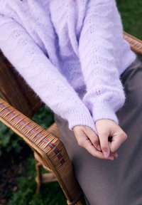 Lila pletena jopica z teksturo, ki spominja na valove. Roke počivajo v naročju, kar razkriva gladko kožo. Vidna je rjava tkanina krila.