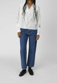 Kvinna klädd i en vit spetsblus, blå raka jeans, svarta strumpor och svarta Mary Jane-skor som står mot en enfärgad bakgrund.