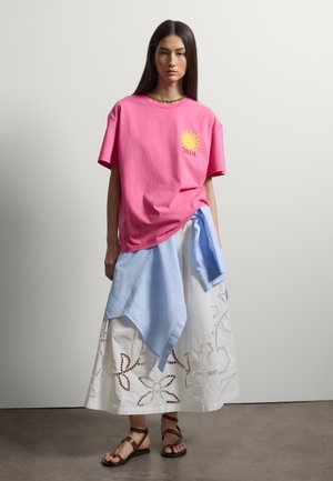 Frau mit langen dunklen Haaren, die ein pinkes T-Shirt mit gelbem Sonnenmotiv, einen weißen bestickten Rock, ein blaues Hemd, das um die Taille gebunden ist, und braune Sandalen trägt.