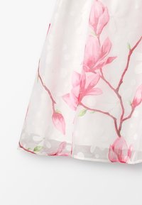 Průhledné šaty s bílým pozadím, zdobené růžovými květy magnólie a zelenými detaily. Tkanina má texturovaný vzor s puntíky.