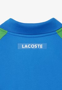 Blå pikétröja med gröna detaljer, med en texturerad tygyta och ruggad krage. Lappen visar "LACOSTE" i vitt på blå bakgrund.