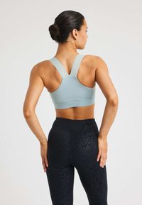 Vrouw met een knot in haar haar, gekleed in een lichtblauwe cross-back sportbeha en zwart met patroon leggings, staat met haar handen op haar heupen en kijkt weg.
