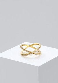 Zlatni prsten s križnim uzorkom i ugrađenim malim dijamantima na bijelom postolju protiv jednostavne sive pozadine.