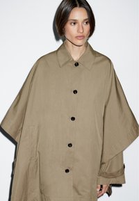 Khaki übergroßer Mantel mit einem Camp-Kragen, schwarzen Knöpfen und seitlichen Taschen. Hergestellt aus leichtem Stoff mit einer glatten Textur.