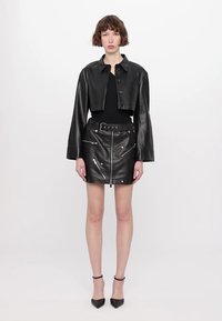Črna usnjena krajšana jakna z dolgimi rokavi, v kombinaciji s črno usnjeno minikrilo z zadrgama in pasom, oblečeno z črnimi petkami.