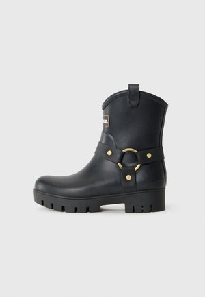 Black mid-calf rubber boot with a thick lug sole, gold ring buckle detail on the side, and a small logo patch near the top.