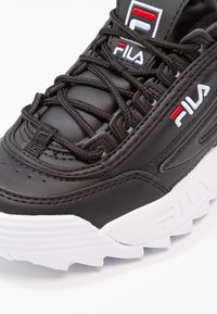 Zapatilla de cuero negra con parte superior texturizada, que cuenta con gruesos cordones negros y una suela de goma blanca. Logo notable de Fila en acentos rojos y blancos.