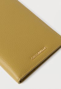 Portofel Coccinelle din piele texturată de culoare muștar. Dispune de detalii aurii cu logo și margini curate pentru un design elegant.